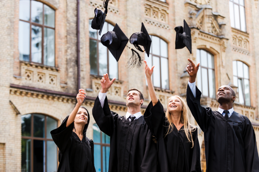 We are finally graduated! Four happy college graduates in graduation gowns throwing their mortar boards and smiling while standing near university